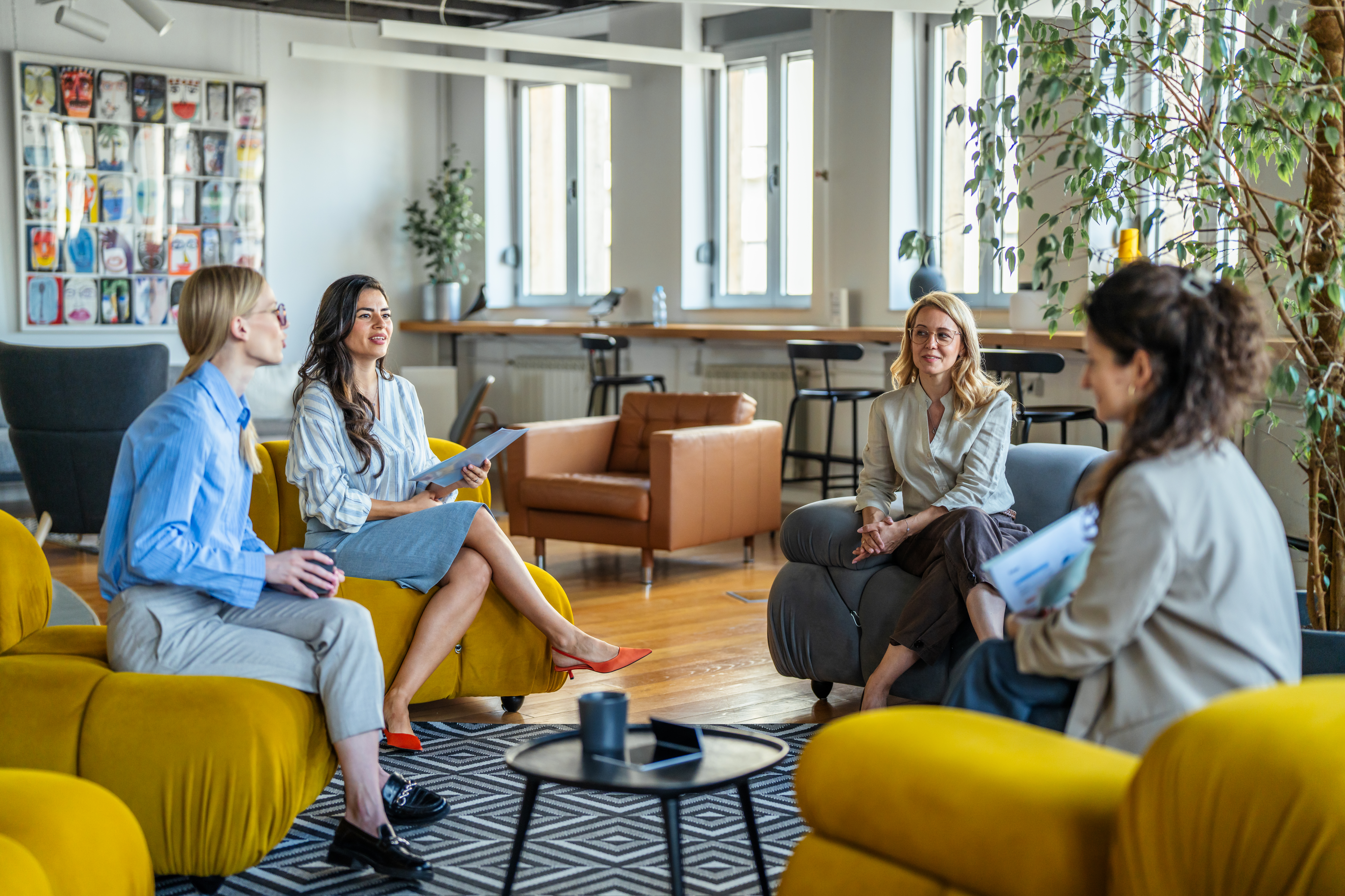 Four businesswomen engaging in a discussion and creative collaboration during a casual meeting in a bright, modern office lounge area